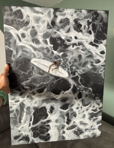 Un tableau représentant une mer agitée vue de dessus avec une surfeuse accrochée à sa planche. Le tout en noir et blanc