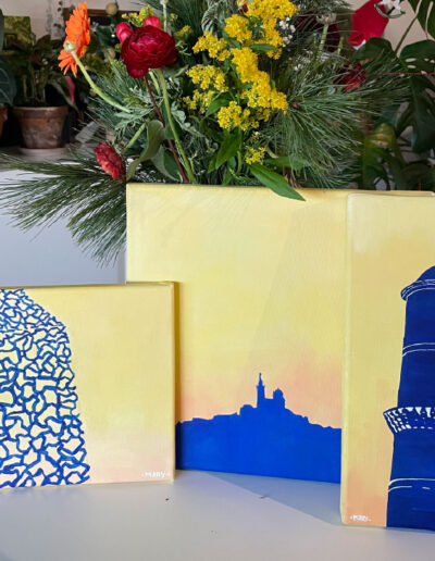 Un tryptique de tableaux représentant des monuments de Marseille, bleus sur fond aux couleurs du coucher de soleil